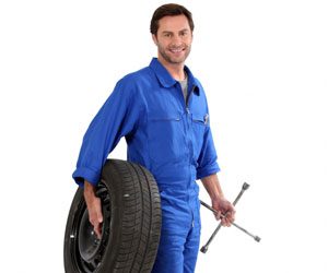 Smiling mechanic in blue coveralls holds a tire and lug wrench against a white background.