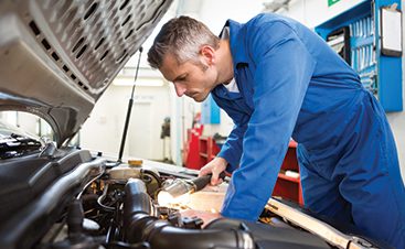Mechanic in blue coveralls leans over an open car hood, inspecting the engine in a repair shop.