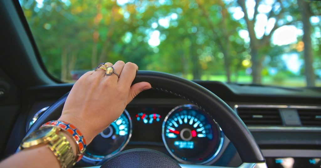 Hand holding a steering wheel while driving along a tree-lined street.