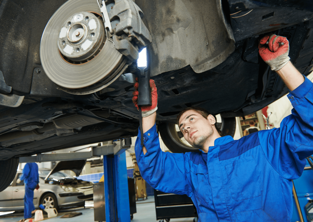 Auto mechanic in blue uniform inspects the underside of a lifted car with a flashlight in a repair shop.