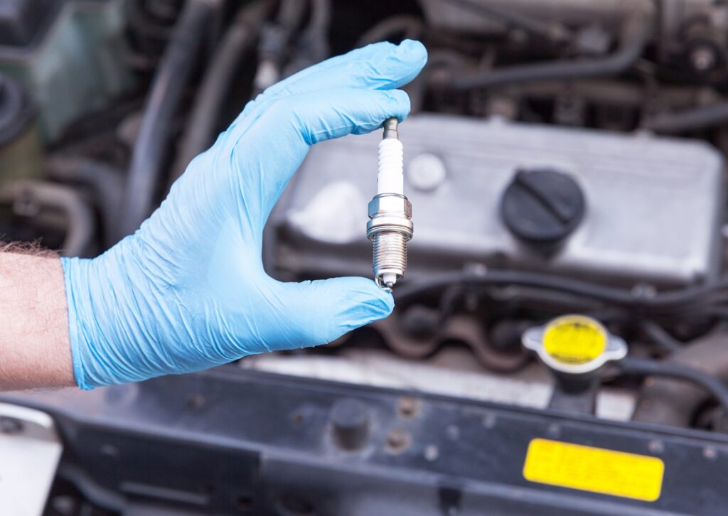 Mechanic wearing blue gloves holding a new spark plug in front of a car engine during a tune-up or repair.