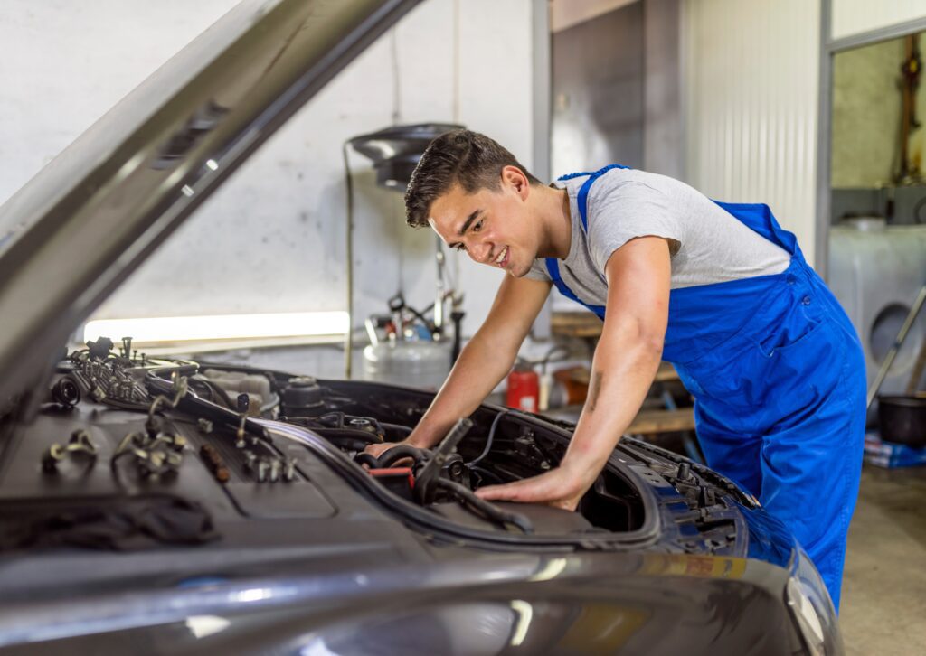 Smiling mechanic in blue overalls working under the hood of a car in a well-lit garage with tools nearby.