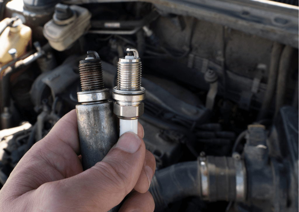 Hand holding a worn spark plug next to a new one, with a car engine in the background.