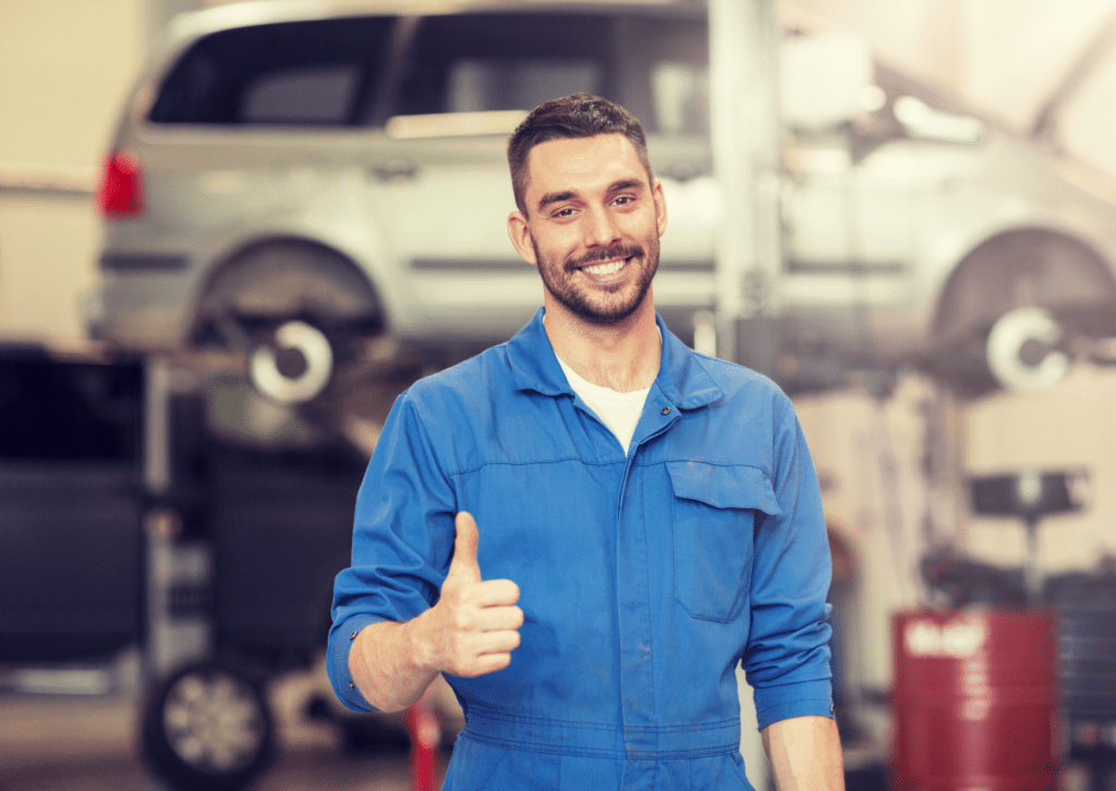Smiling auto mechanic in a blue uniform giving a thumbs-up in a repair shop with a car on a lift.