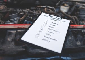 Car maintenance checklist on a clipboard resting on an open engine bay, listing essential vehicle service tasks.