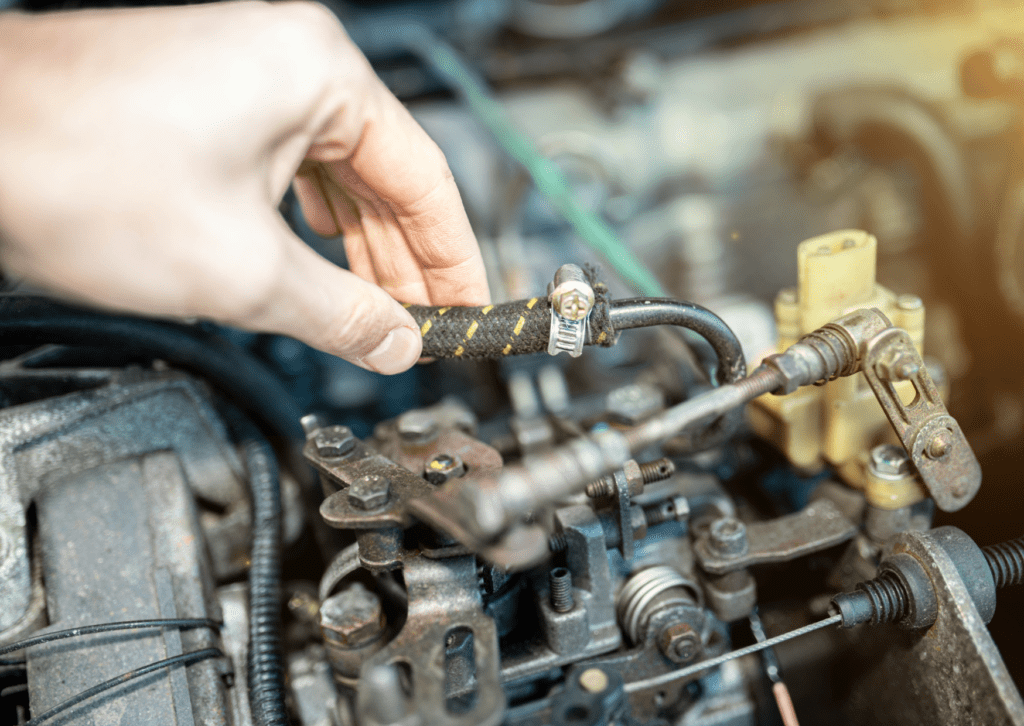 Hand inspecting a worn fuel hose with a metal clamp in an old car engine bay.