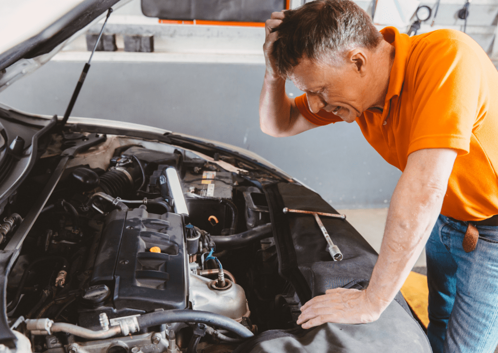 Frustrated man in an orange shirt inspecting a car engine with tools and a work light in a garage.