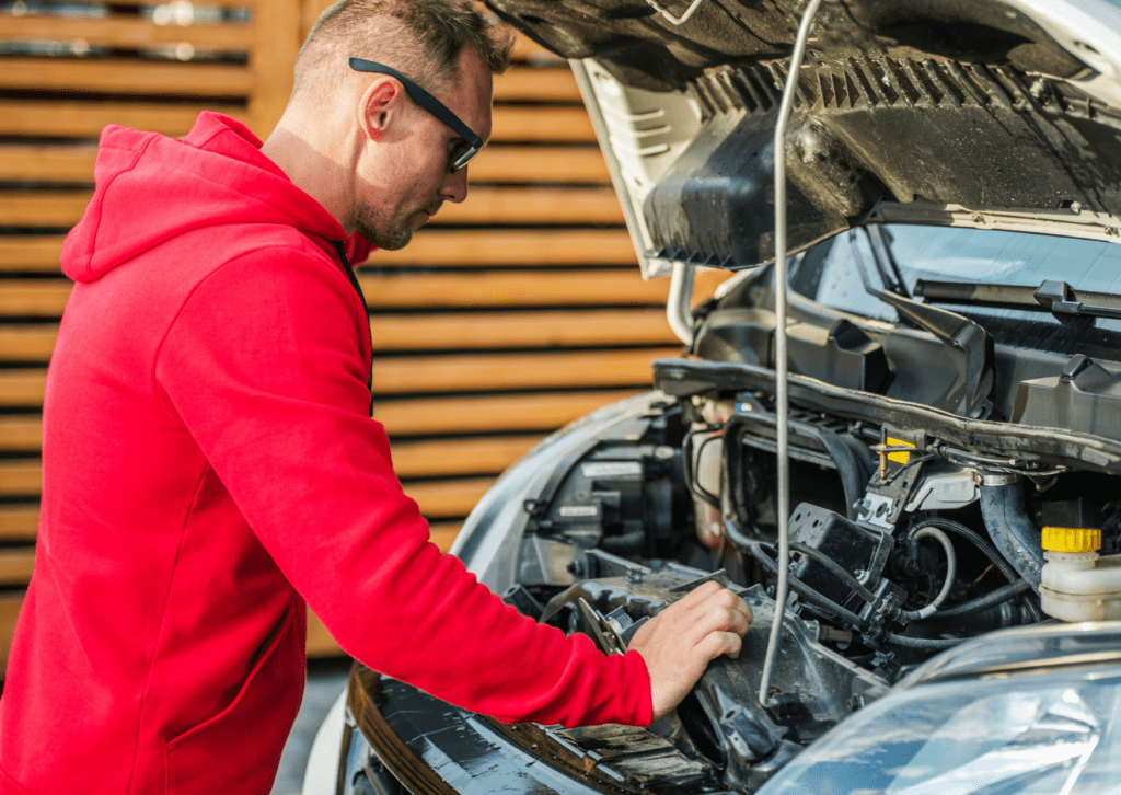 Man in a red hoodie inspecting a car engine with the hood open, checking for maintenance or repairs.