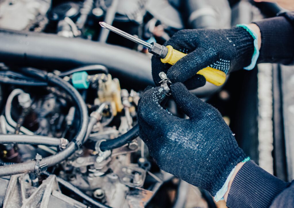 Mechanic in gloves adjusts hose clamp with screwdriver on engine bay during diagnosis of fuel system issues and leaks.