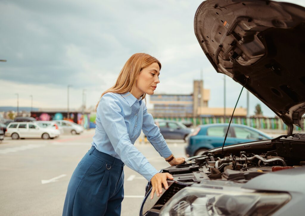 Woman inspecting open car hood in parking lot, leaning over engine bay under cloudy daylight sky.