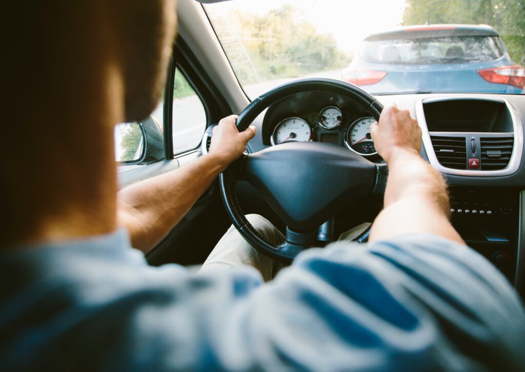 Driver holding steering wheel in traffic, viewed from back seat, with dashboard and ahead visible.