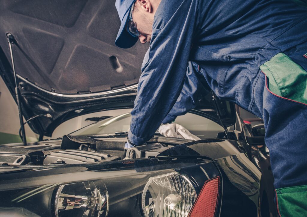 Mechanic working under open hood, inspecting engine bay during auto repair in a service garage.