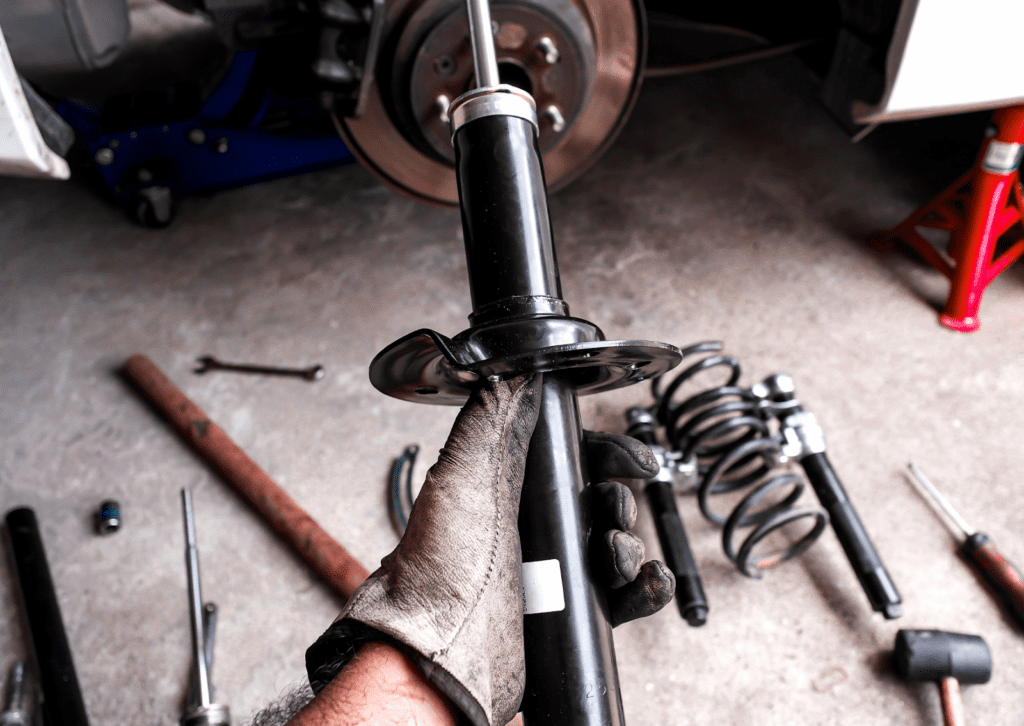 Gloved mechanic holds new shock absorber during suspension repair beside removed brake assembly.