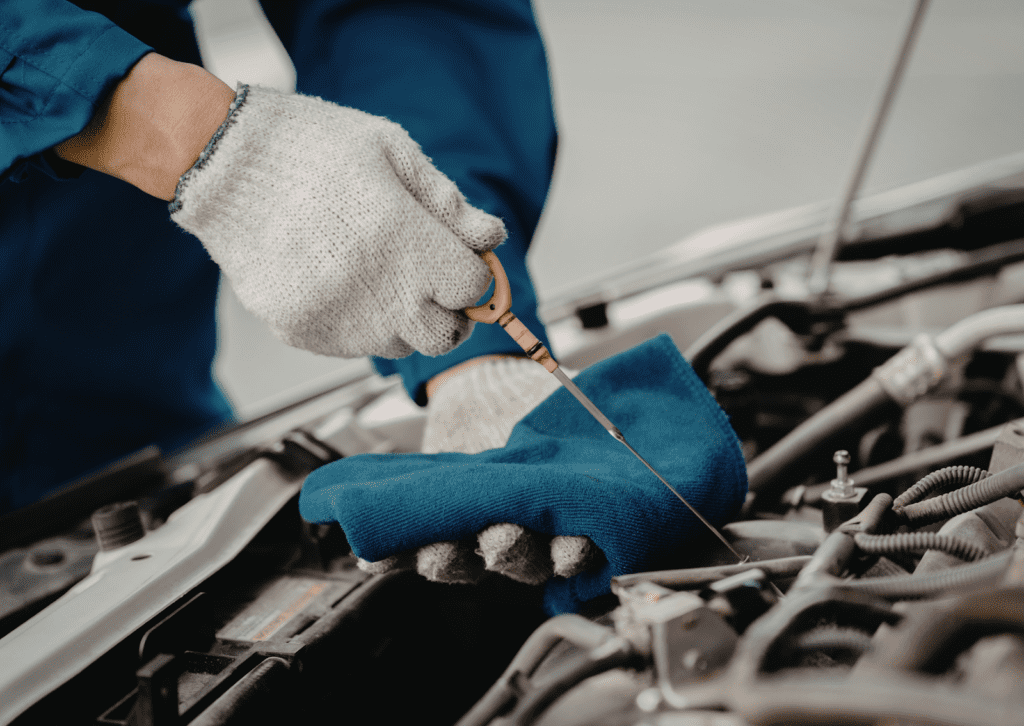 Gloved mechanic checks dipstick under hood, showing what causes oil leaks in car inspection.