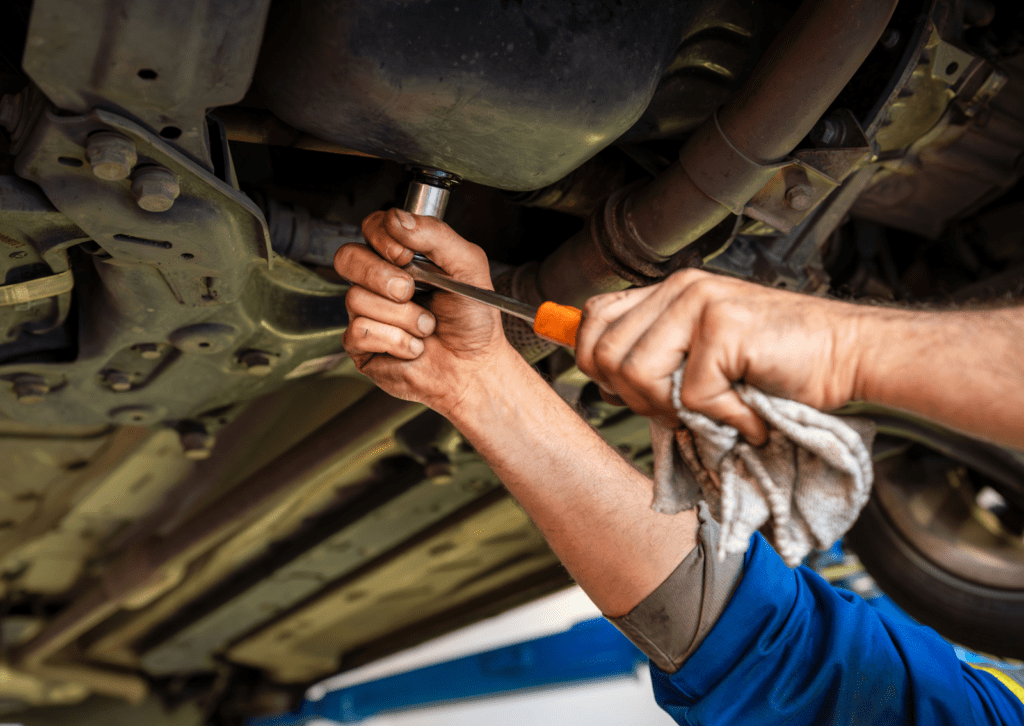 Mechanic loosens oil pan bolt under vehicle, illustrating what causes oil leaks in car damage.