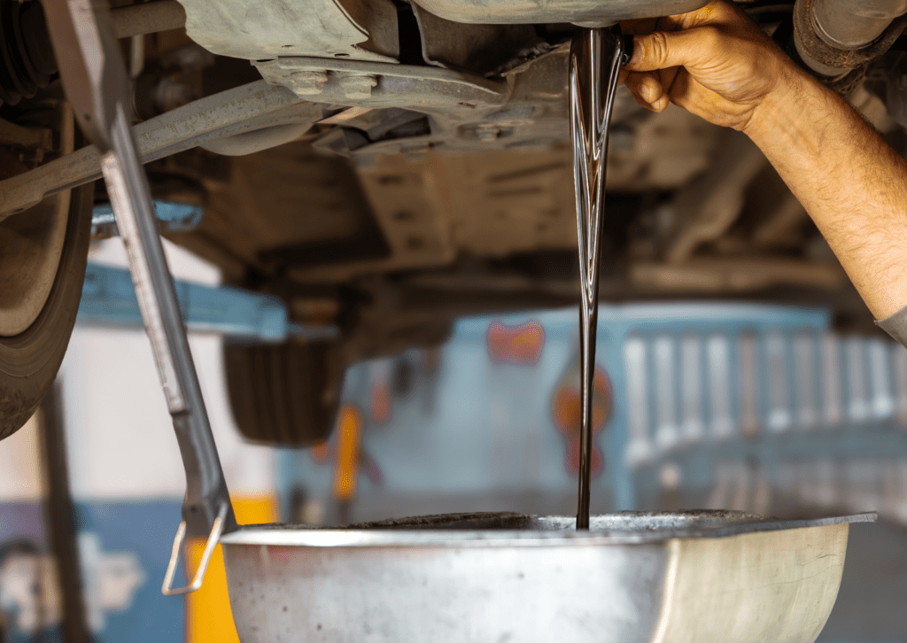 Mechanic drains engine oil into pan beneath lifted car during routine maintenance service check.
