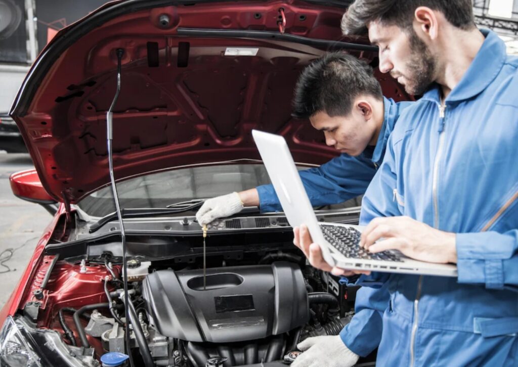 Two mechanics inspecting car engine with laptop and dipstick under open hood in auto service bay.
