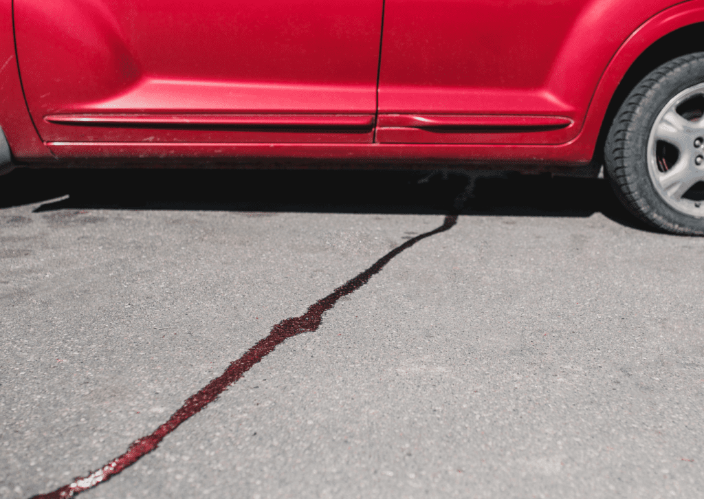 Red car parked on pavement with a long dark fluid trail leaking from underneath the vehicle 