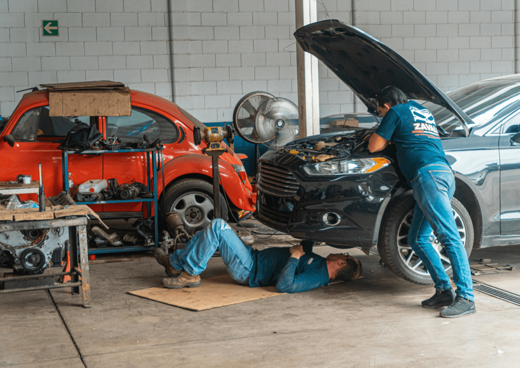 Mechanics inspect a car with the hood open while one works underneath in a busy repair shop 