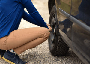 Driver checking a wheel after hearing a car clunking noise on a gravel roadside beside vehicle 