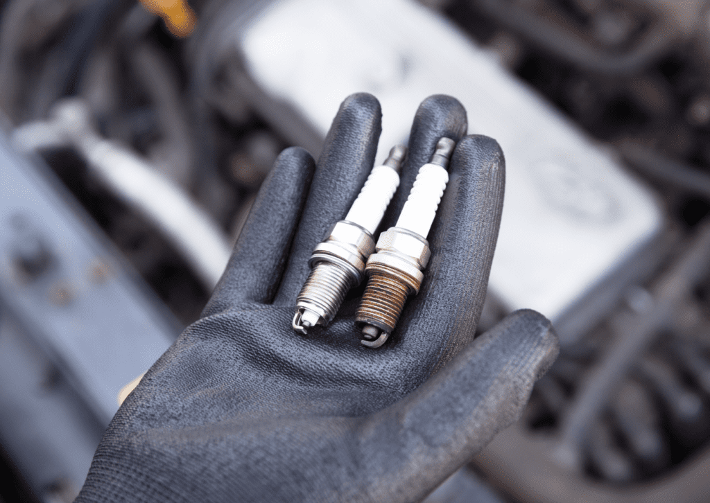 Gloved hand holding old and new spark plugs over engine bay during ignition system service 