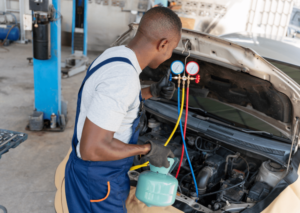 Mechanic servicing car AC system with manifold gauges and refrigerant tank under open hood