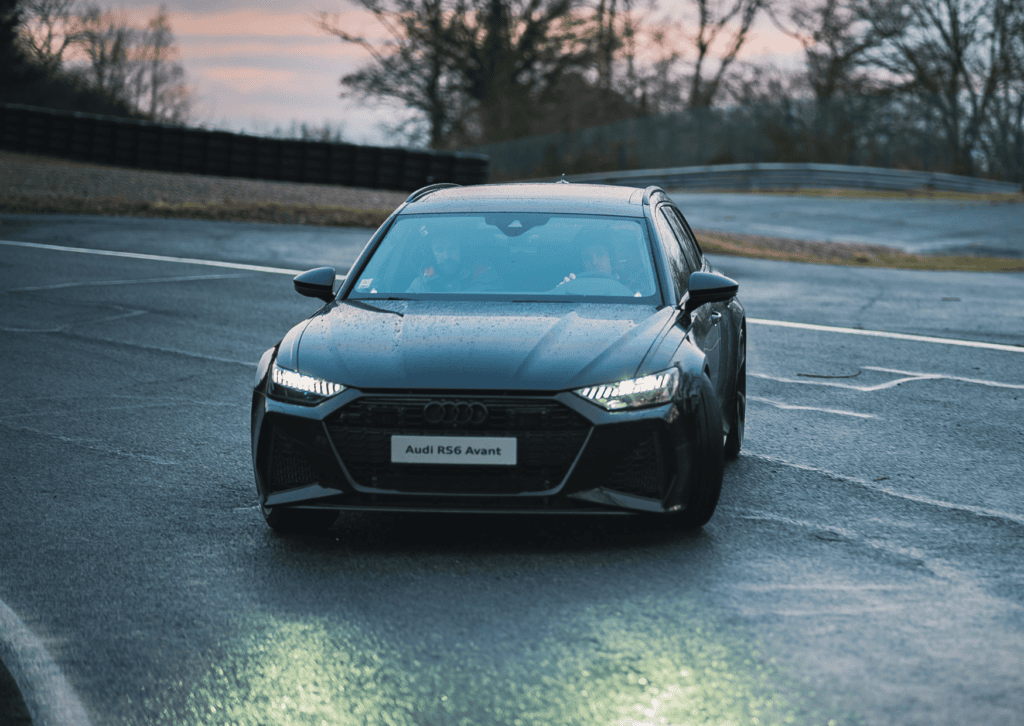 Black Audi RS6 Avant driving across a wet racetrack with bright headlights under a cloudy sky.