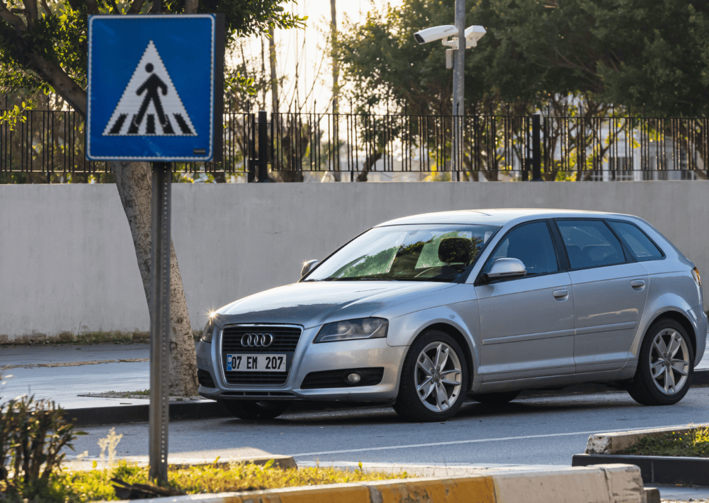 Silver Audi hatchback parked beside a pedestrian crossing sign on a quiet, tree-lined street.