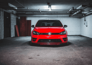 Front view of a red Volkswagen parked inside a dim concrete garage beneath exposed ceiling pipes.
