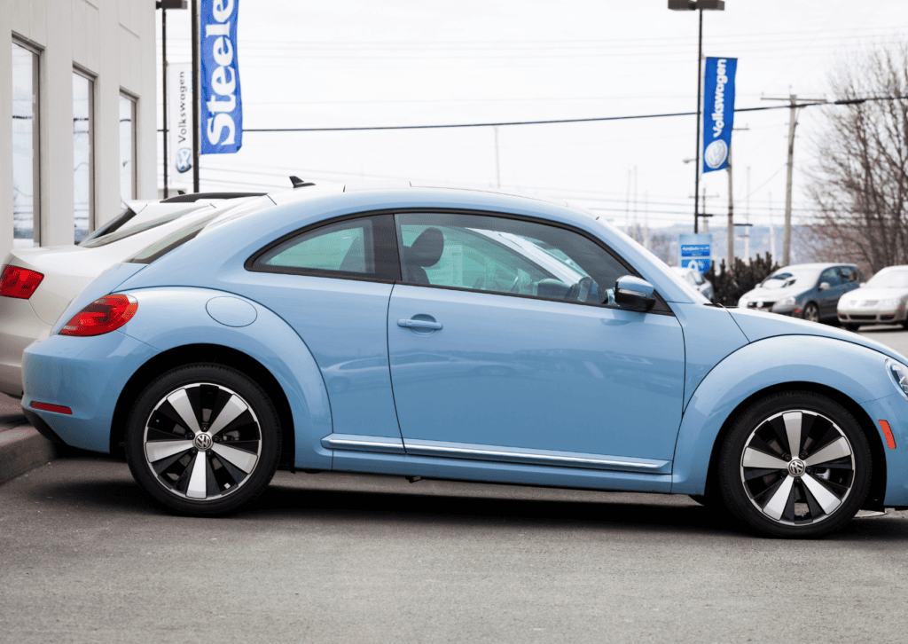 Light blue Volkswagen Beetle parked outside a dealership beside other vehicles and branded flags.