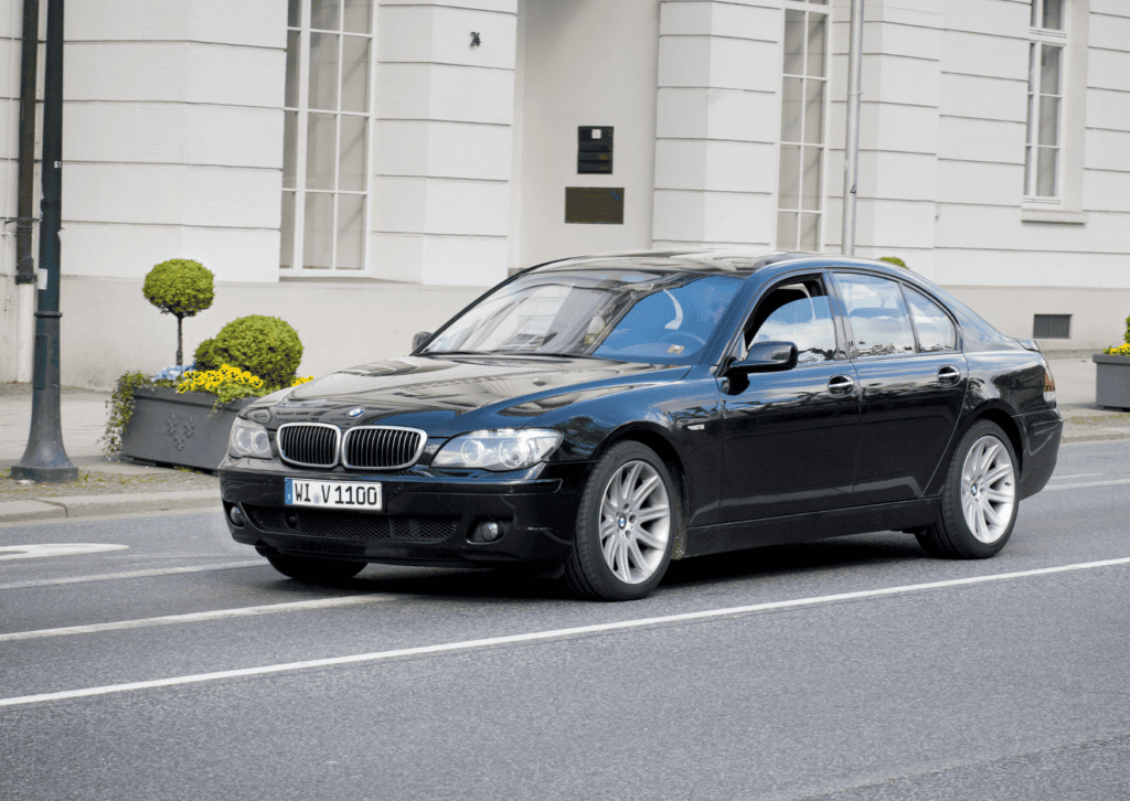 Black BMW luxury sedan driving past a white building with trimmed shrubs and flower planters.