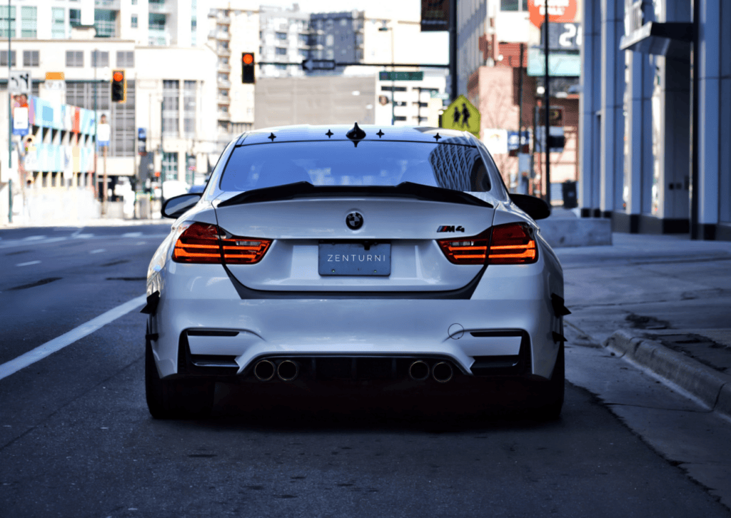 Rear view of a white BMW M4 with illuminated taillights parked on a quiet downtown street.