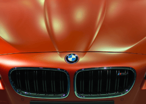 Close-up of a BMW emblem and twin kidney grille on a metallic orange car under showroom lights.
