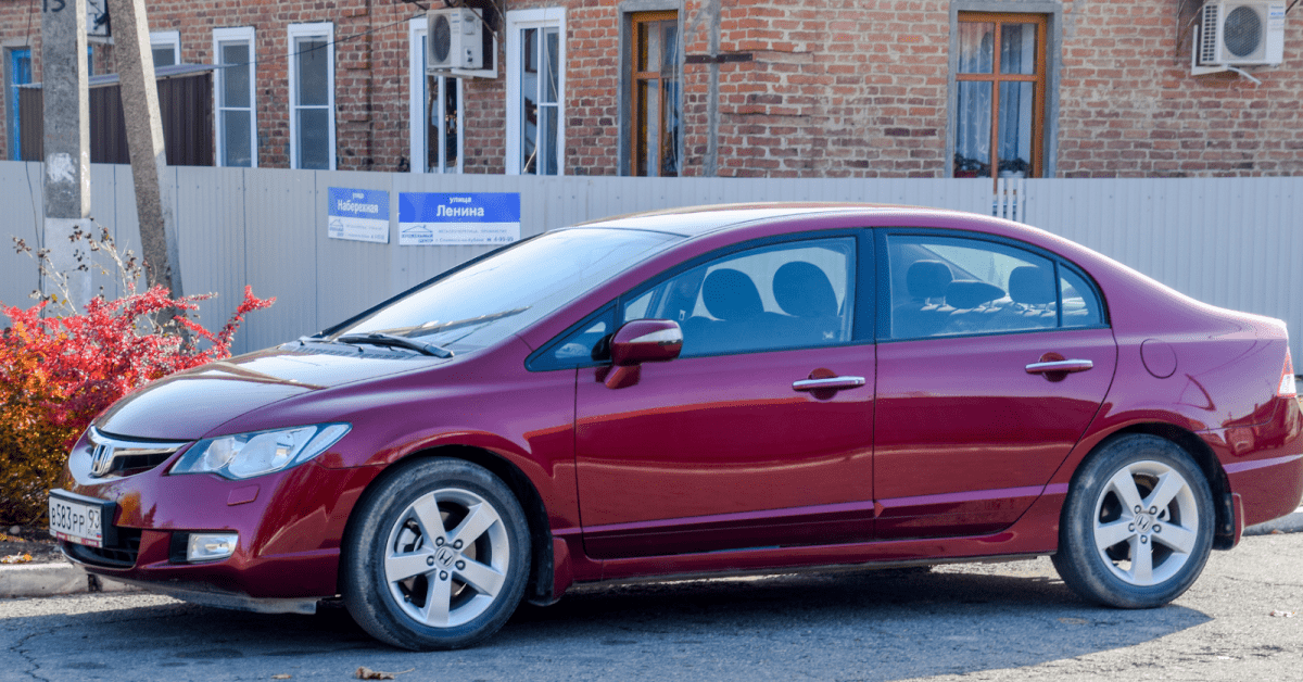 Maroon Honda Civic sedan with alloy wheels parked on sunlit street before brick building and grey fence.