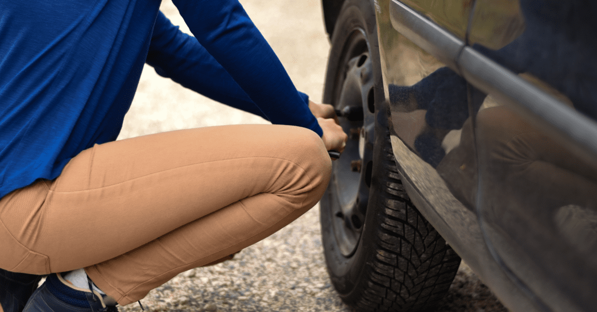 Driver checking a wheel after hearing a car clunking noise on a gravel roadside beside vehicle 