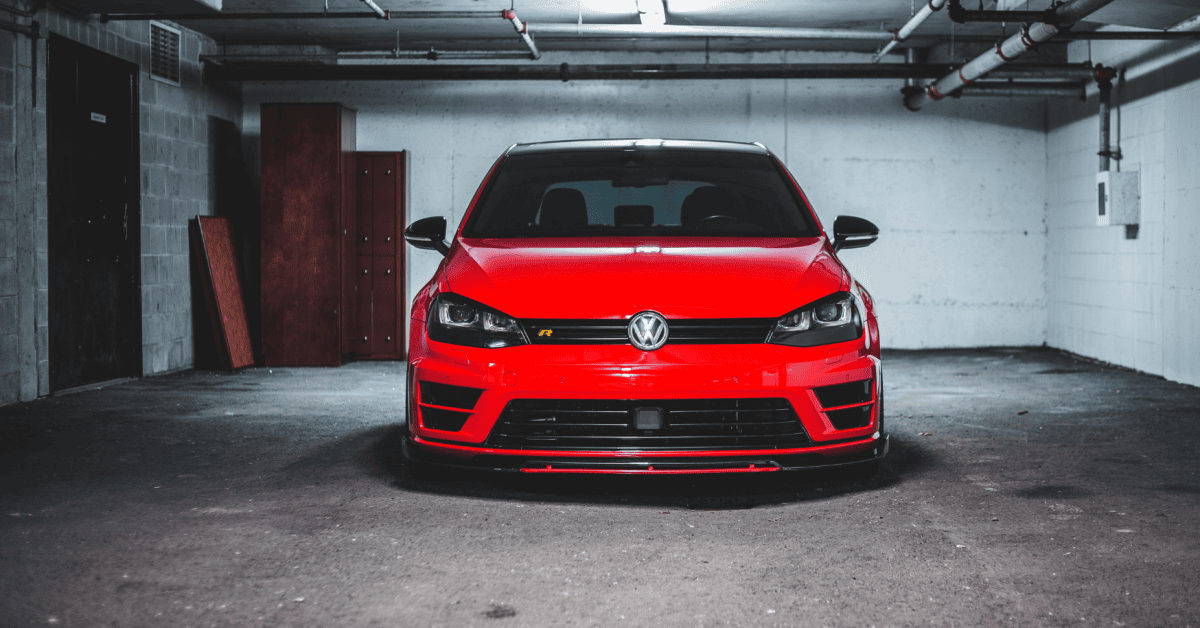 Front view of a red Volkswagen parked inside a dim concrete garage beneath exposed ceiling pipes. 