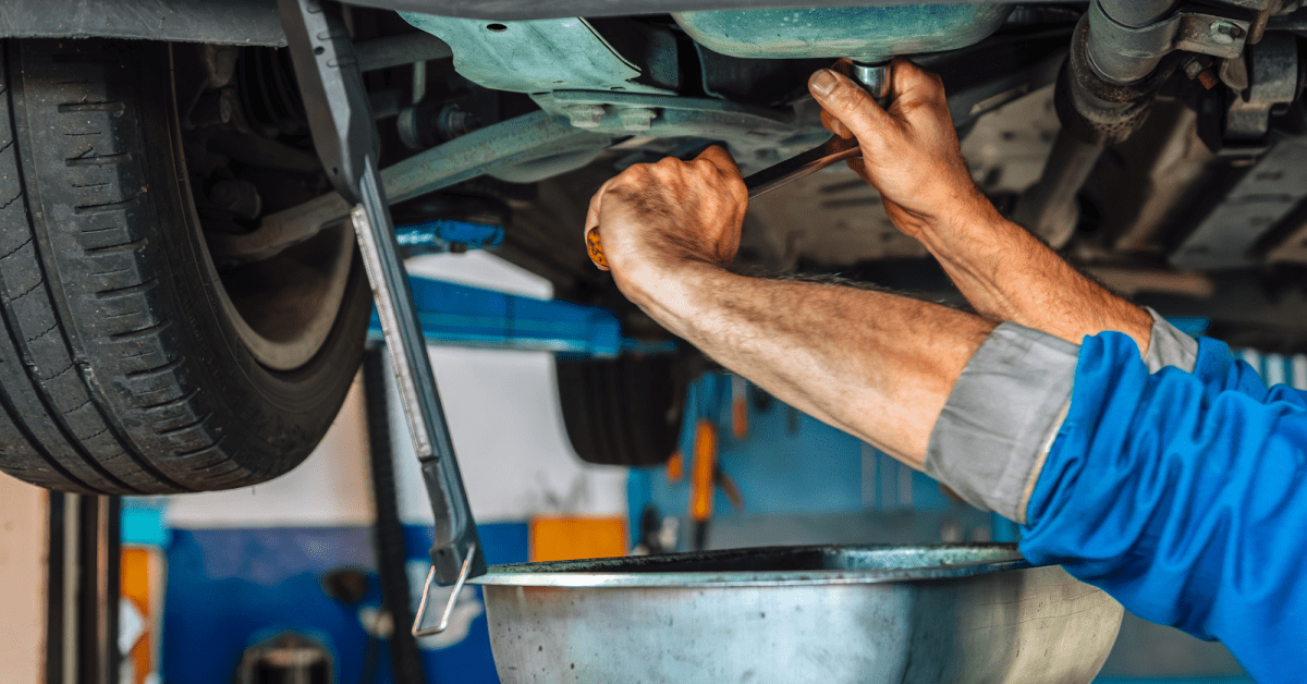 Mechanic loosens drain plug under lifted car, showing what causes oil leaks in car during service.
