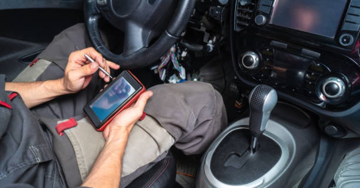 Diagnostics of car failures: an electrician in gray clothes is sitting in the car's interior and reading the trouble codes from a portable auto scanner with OBD2 interface. Close-up of hands.