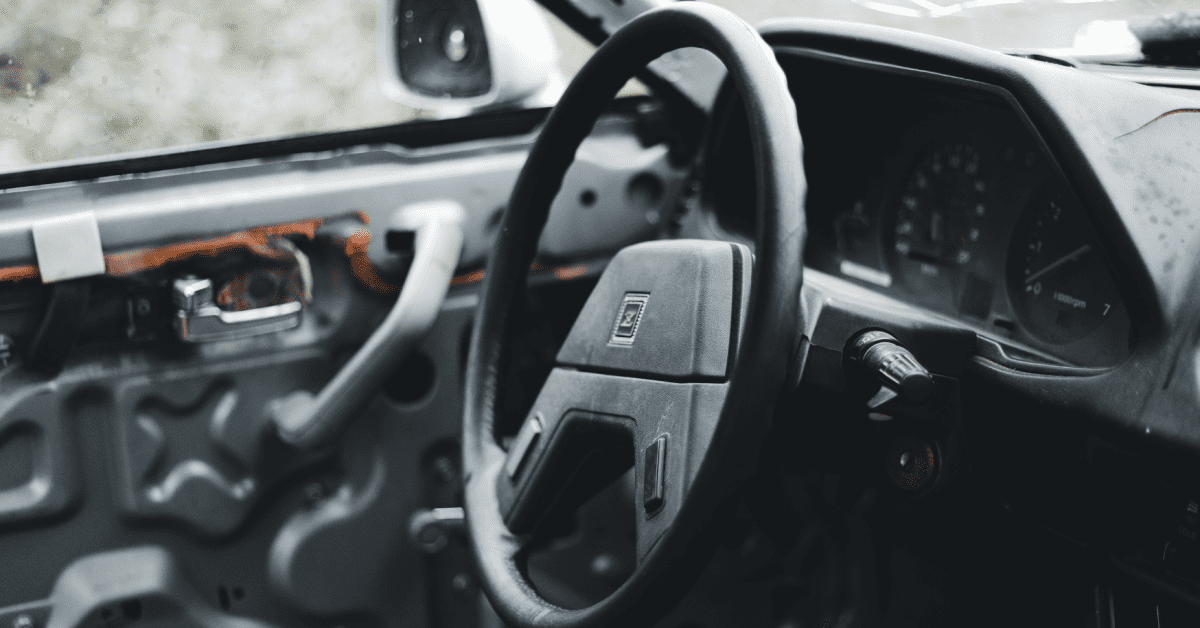 Close-up of an old car interior with a worn-out steering wheel and exposed door panel, showing steering and suspension wear.